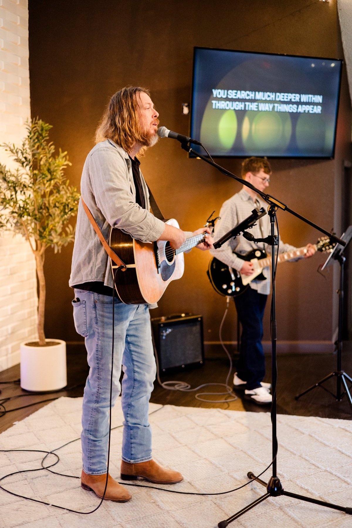 A worship leader sings and plays acoustic guitar beside a guitarist, with the song lyrics on a screen behind them.