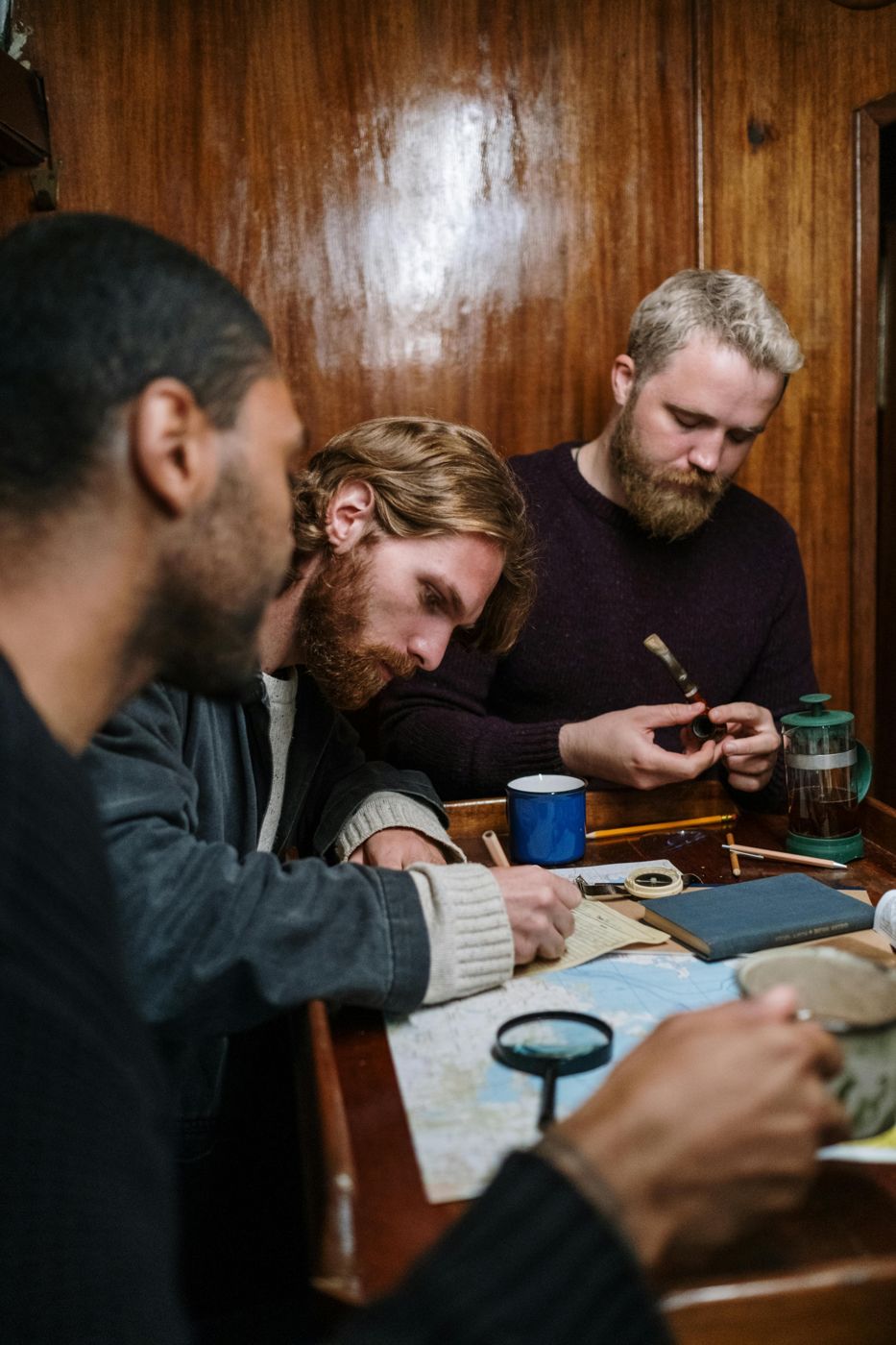 Three men gathered around a wooden table with notebooks and a map, studying together